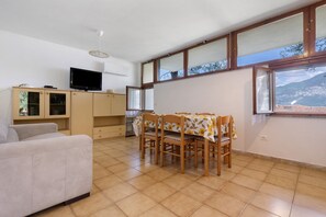 A living room with a sofa, a dining table with chairs, a TV on a cabinet, and large windows with a mountain view.