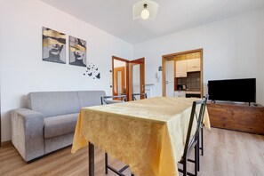 Un salon avec un canapé, une table à manger recouverte d’une nappe, un téléviseur et un coin cuisine visible à travers une porte ouverte.
