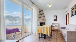 A living room with a sofa, a dining table with chairs, a fireplace, and a view of the sea.
