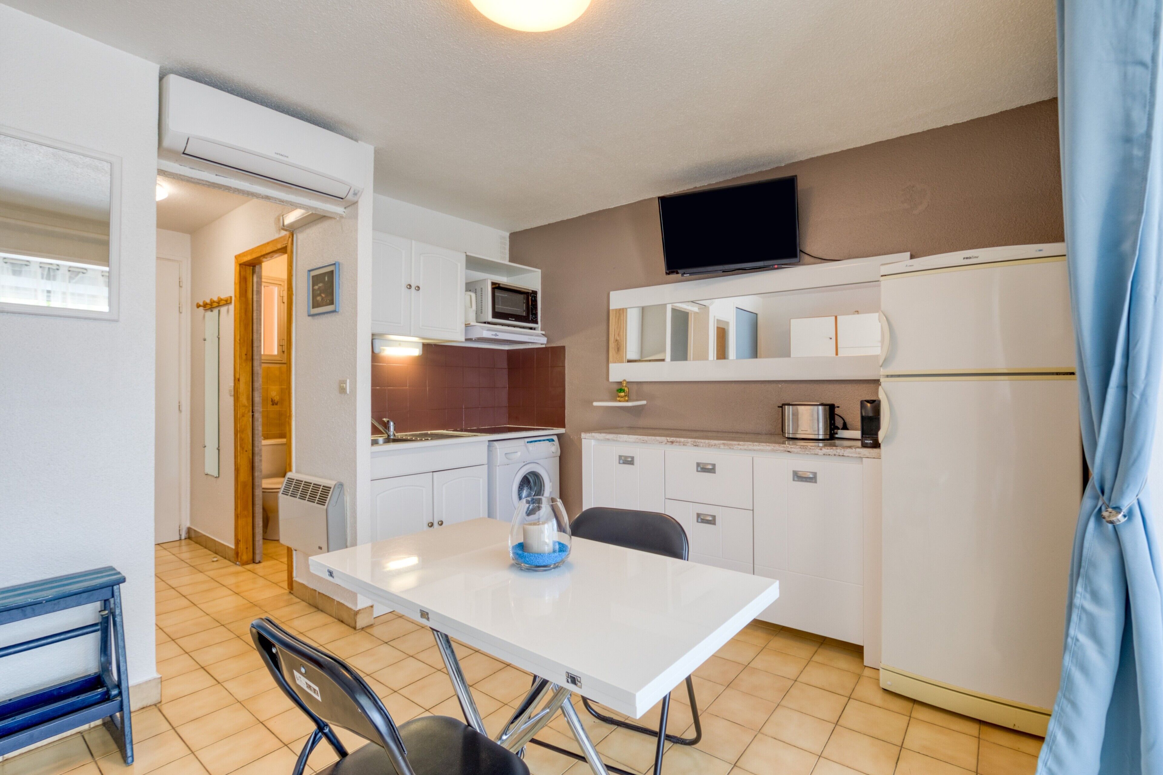 A compact kitchen with a dining table, chairs, a microwave, a coffee maker, and a TV mounted above the counter.