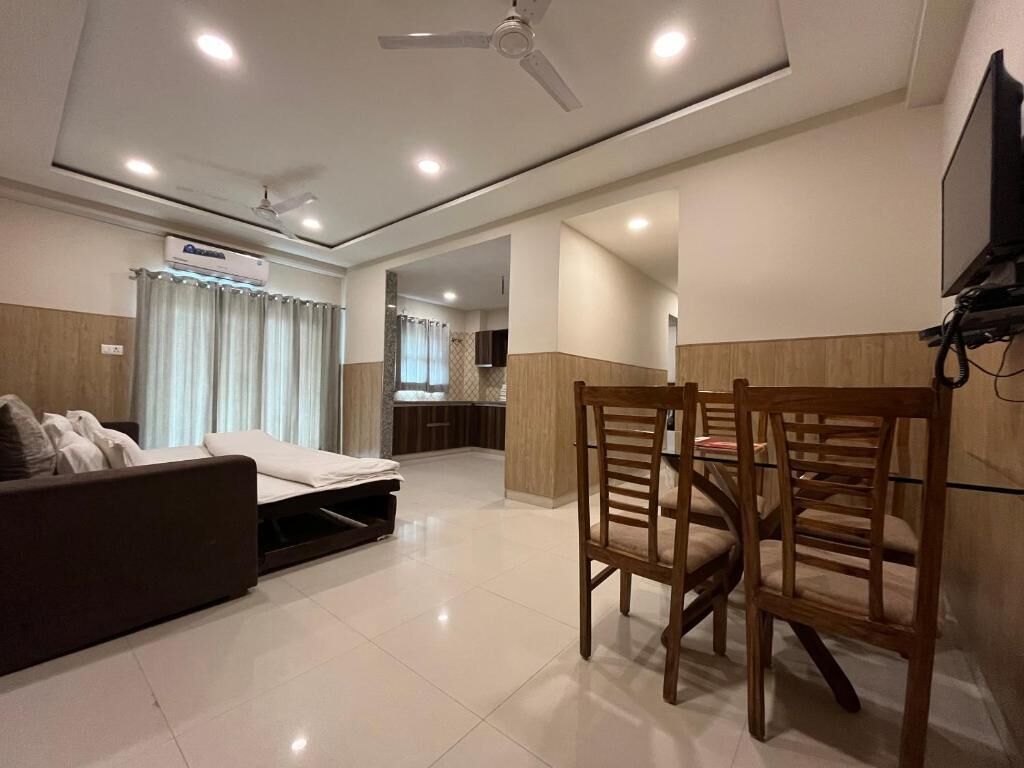 A modern living room with a sofa, a dining table with chairs, a ceiling fan, recessed lighting, and a kitchen area in the background.