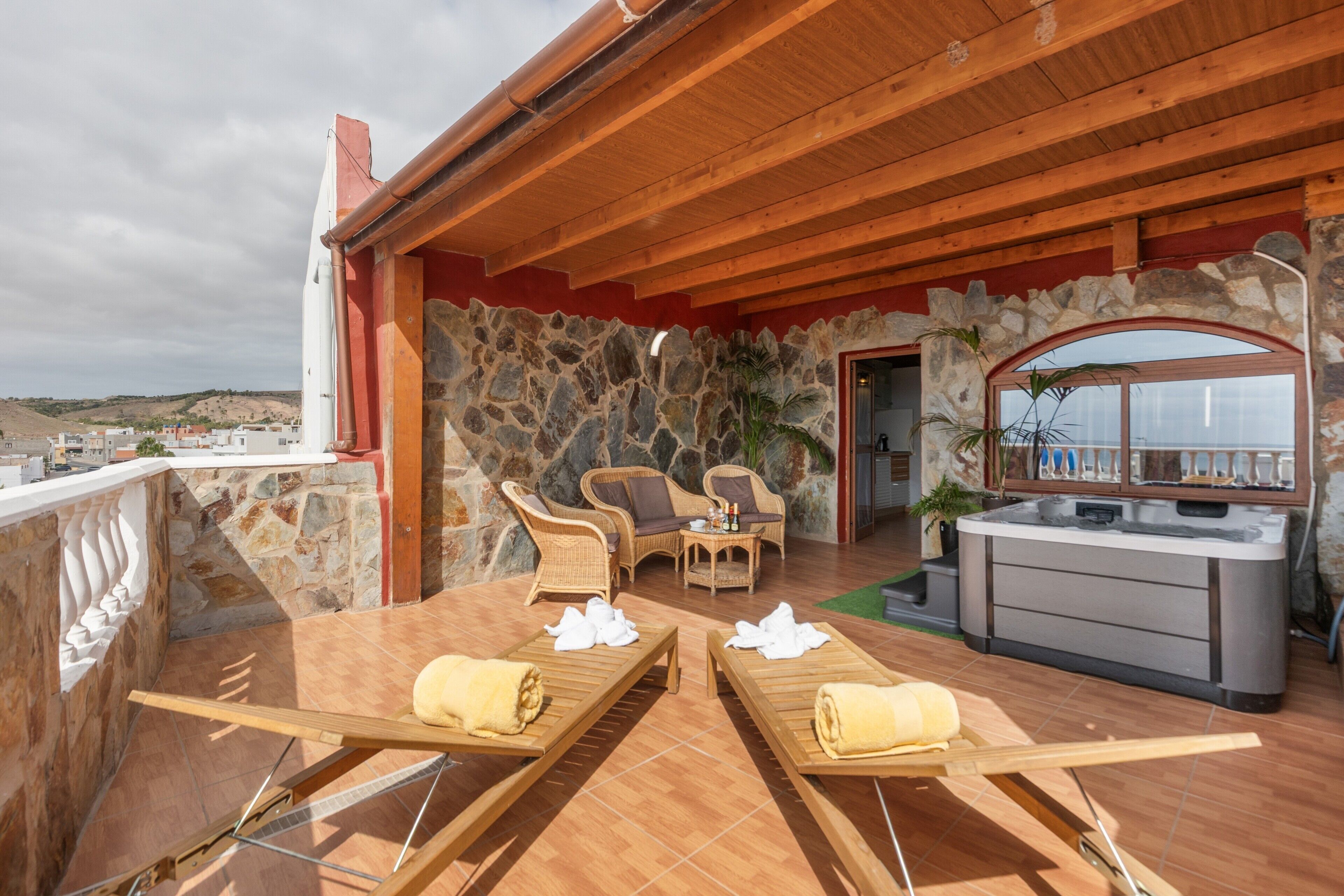 Una terraza con tumbonas de madera, un muro de piedra, muebles de mimbre y un jacuzzi.