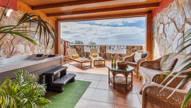 A sunlit outdoor lounge area with stone walls, a hot tub, wicker furniture, and a view of the sky. - Chabela's Penthouse Apartment with Sea Views, Jacuzzi, and Air Conditioning (La Lajita)
