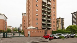 A multi-story residential building with balconies, a gated entrance, and a parking area with cars.