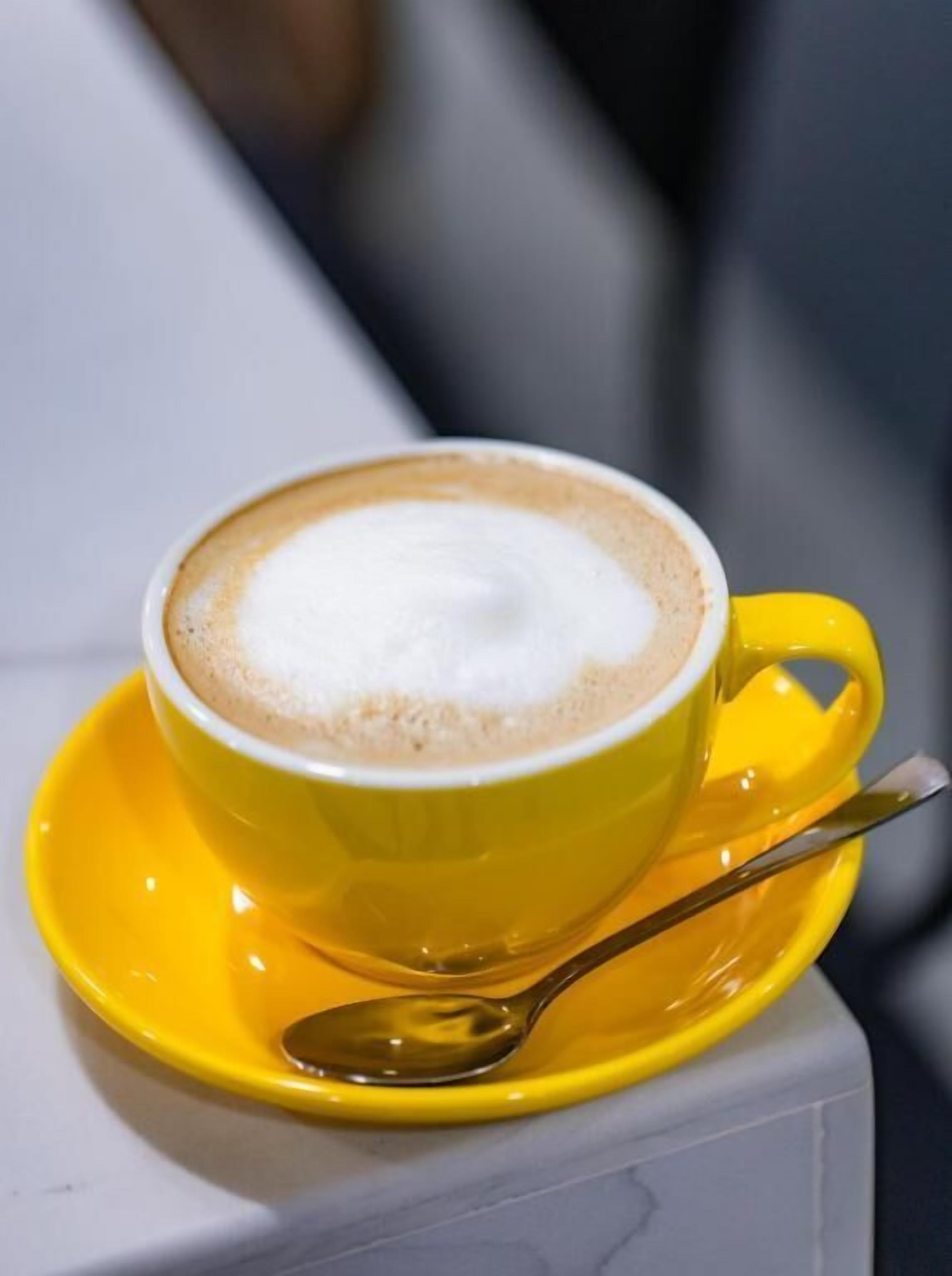 A yellow cup of coffee with a spoon on a saucer, placed on a white surface.