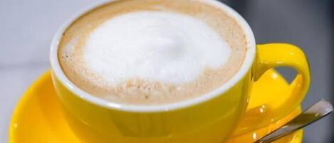 A yellow cup of coffee with a spoon on a saucer, placed on a white surface.