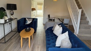 Un salon moderne avec un canapé bleu, une table basse en bois et un téléviseur posé sur un meuble noir. On aperçoit un escalier avec une rampe blanche à l’arrière-plan.