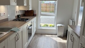 Une cuisine moderne avec des meubles de couleur bois clair, une cuisinière à gaz et une grande fenêtre donnant sur une clôture en bois et une allée.