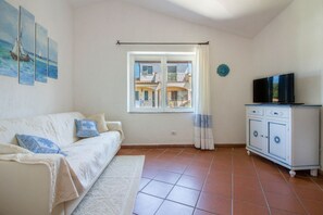 A room with a sofa, a TV on a cabinet, a window with a view of buildings, and a wall with paintings.