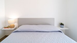 Een net opgemaakt bed met een wit hoofdbord, een bedlampje, een klein plankje met boeken en een plant, en een witte nachtkastje.