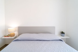 Ein ordentlich bezogenes Bett mit weißem Kopfteil, einer Nachttischlampe, einer kleinen Ablage mit Büchern und einer Pflanze sowie einem weißen Nachttisch.