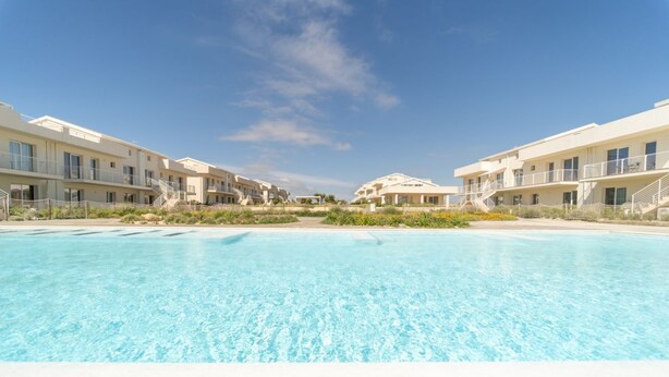A swimming pool with modern buildings, a clear sky, and a sandy beach.