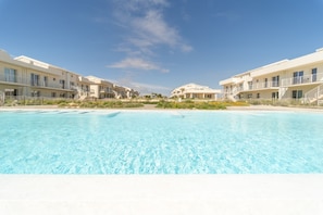 A swimming pool with modern buildings, a clear sky, and a sandy beach.