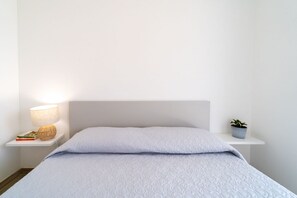 Ein ordentlich bezogenes Bett mit weißem Kopfteil, einer Nachttischlampe, einer kleinen Ablage mit Büchern und einer Pflanze sowie einem weißen Nachttisch.