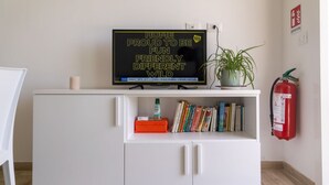 Een witte kast met een televisie bovenop, waarop een bericht staat. Daaronder boeken en een klein oranje doosje. Een potplant staat op de kast, en een brandblusser is aan de rechterkant van de muur bevestigd.