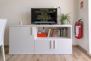 A white cabinet with a television on top displaying a message. Below the TV, there are books and a small orange box. A potted plant is on the cabinet, and a fire extinguisher is mounted on the wall to the right.