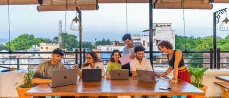 A group of people sitting at a wooden table with laptops, under a covered area with hanging lights, overlooking a cityscape.