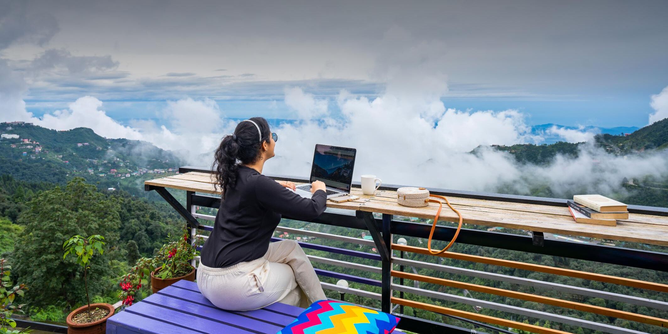 A person working on a laptop at a wooden table with a view of a mountainous landscape.