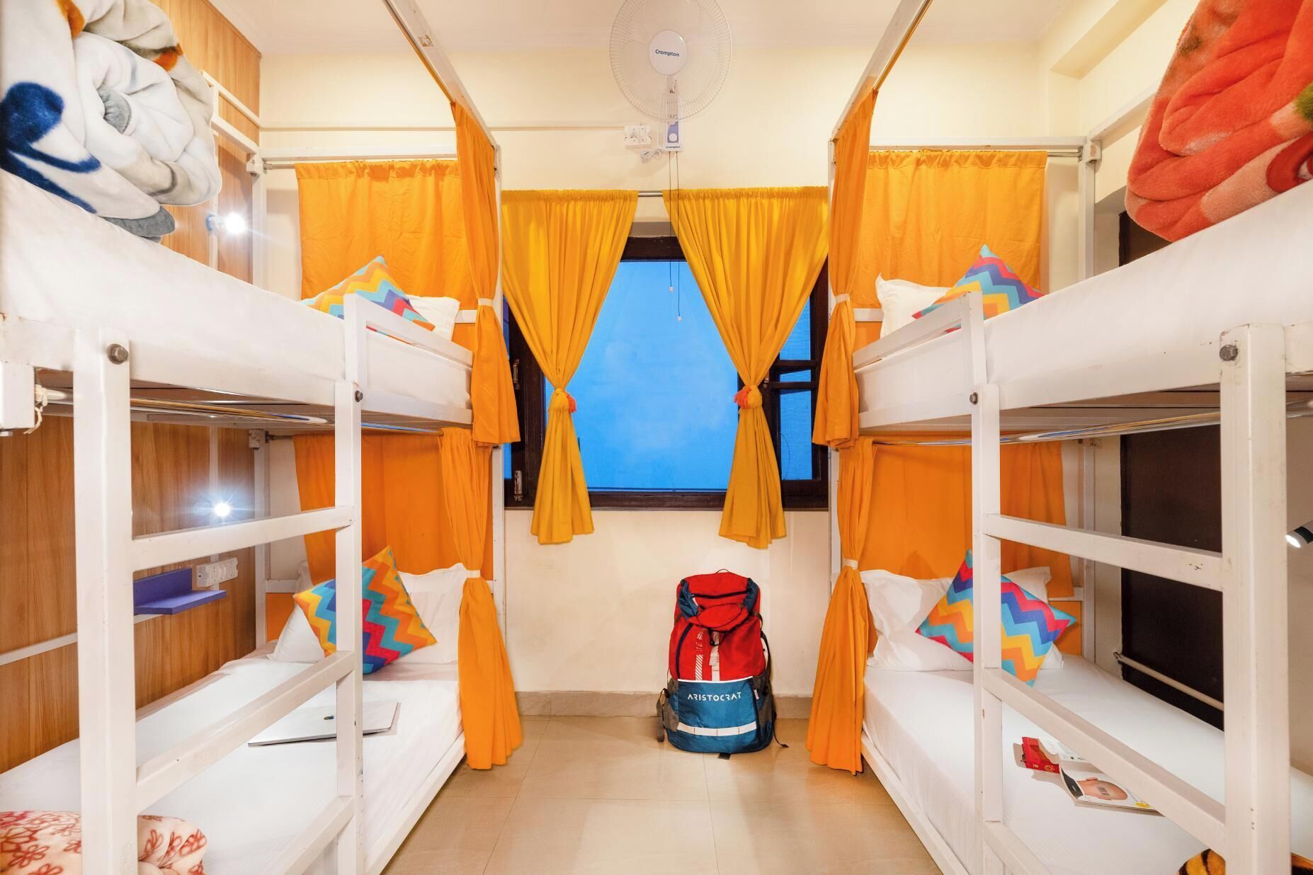 A dormitory room with bunk beds, orange curtains, a fan, and a red backpack on the floor.