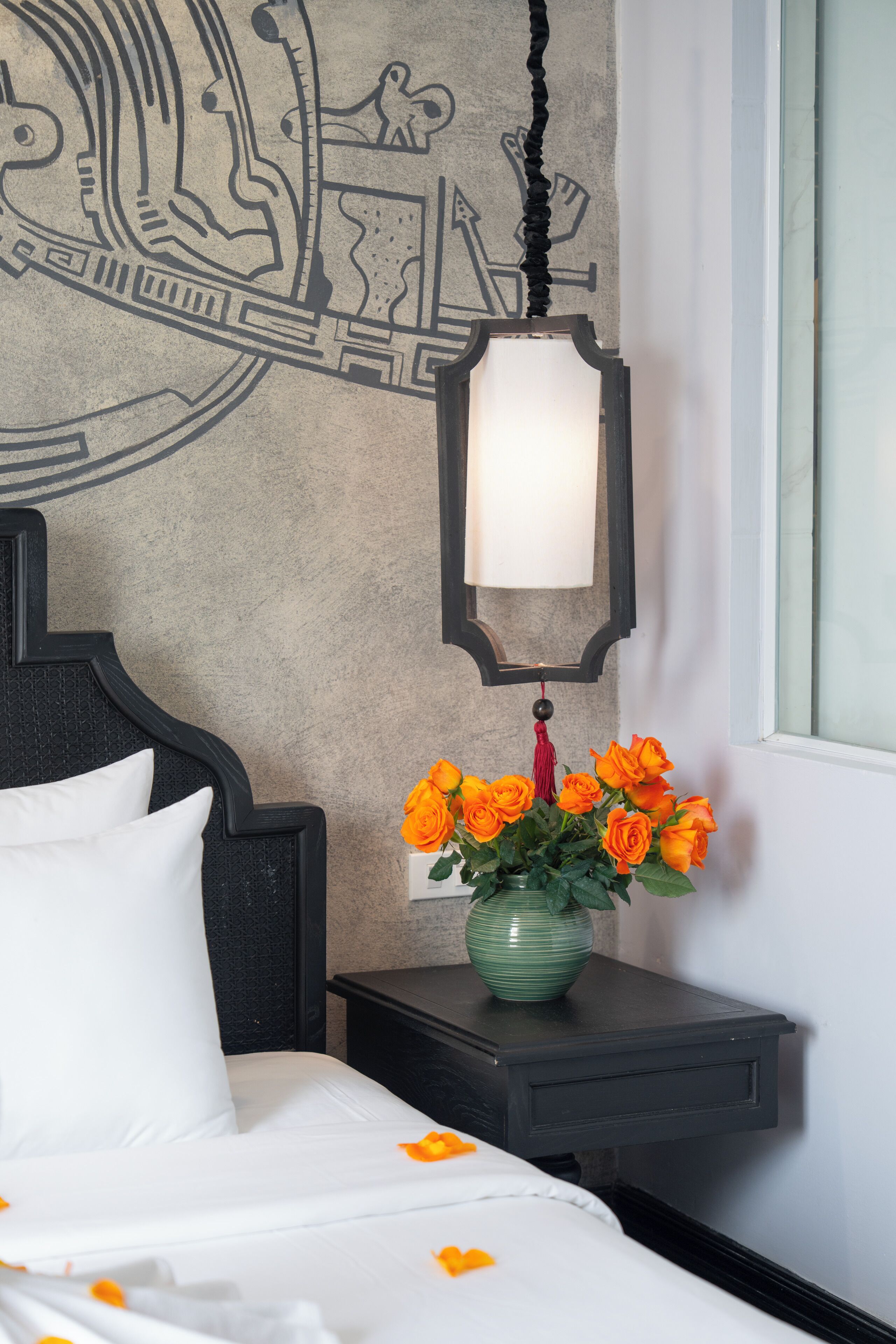 Un lit avec une literie blanche, un vase vert contenant des fleurs orange, un mur orné d’un motif décoratif et un luminaire suspendu.