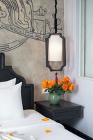 A bed with white bedding, a green vase with orange flowers, a wall with a decorative design, and a hanging light fixture.