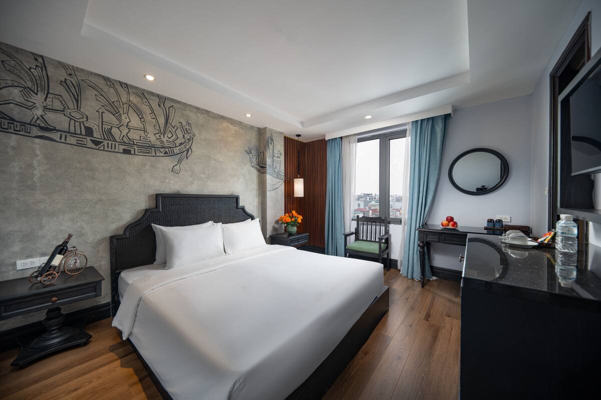 A hotel room with a large bed, a desk with a mirror, a small table with flowers, and a view of the city through the window.
