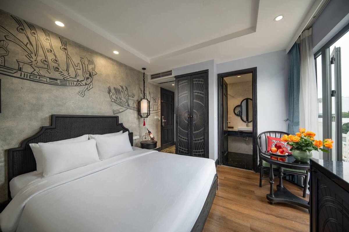 A hotel room with a large bed, a wall mural, a dining area with a table and chairs, and a view through a window.