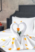 Una cama con una colcha blanca, una toalla dispuesta en forma de corazón y una sola rosa naranja.