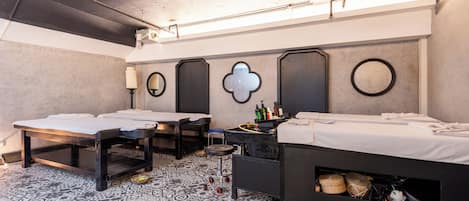 A hotel room with two beds, a small table, a chair, and a mirror. The room has a patterned floor and a ceiling with exposed pipes.