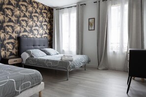 Une chambre moderne avec deux lits, une grande fenêtre avec des rideaux légers et un mur orné d’un motif de feuilles tropicales.