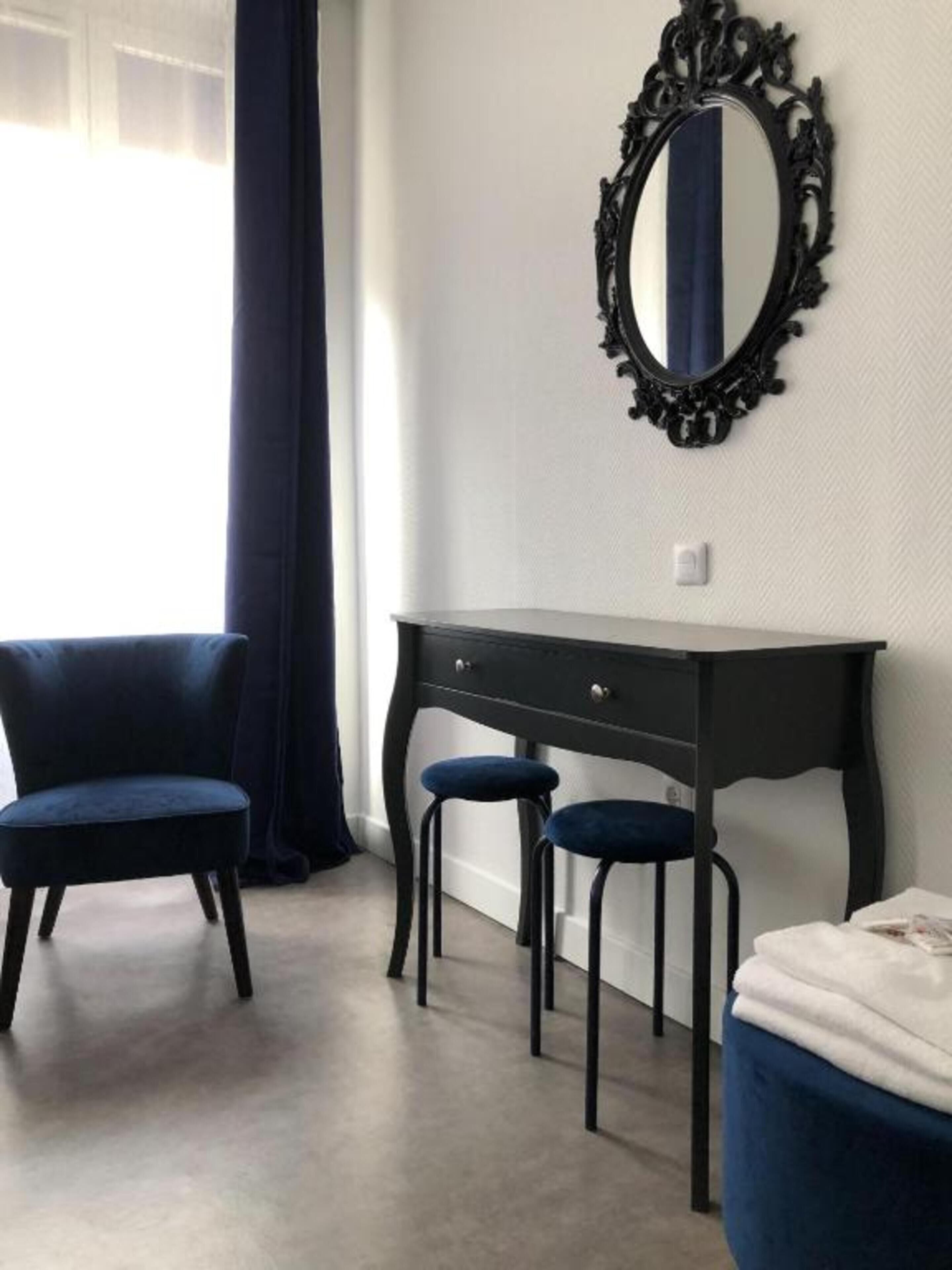 A room with a dark wooden desk, two blue chairs, a large mirror, and a white bed.