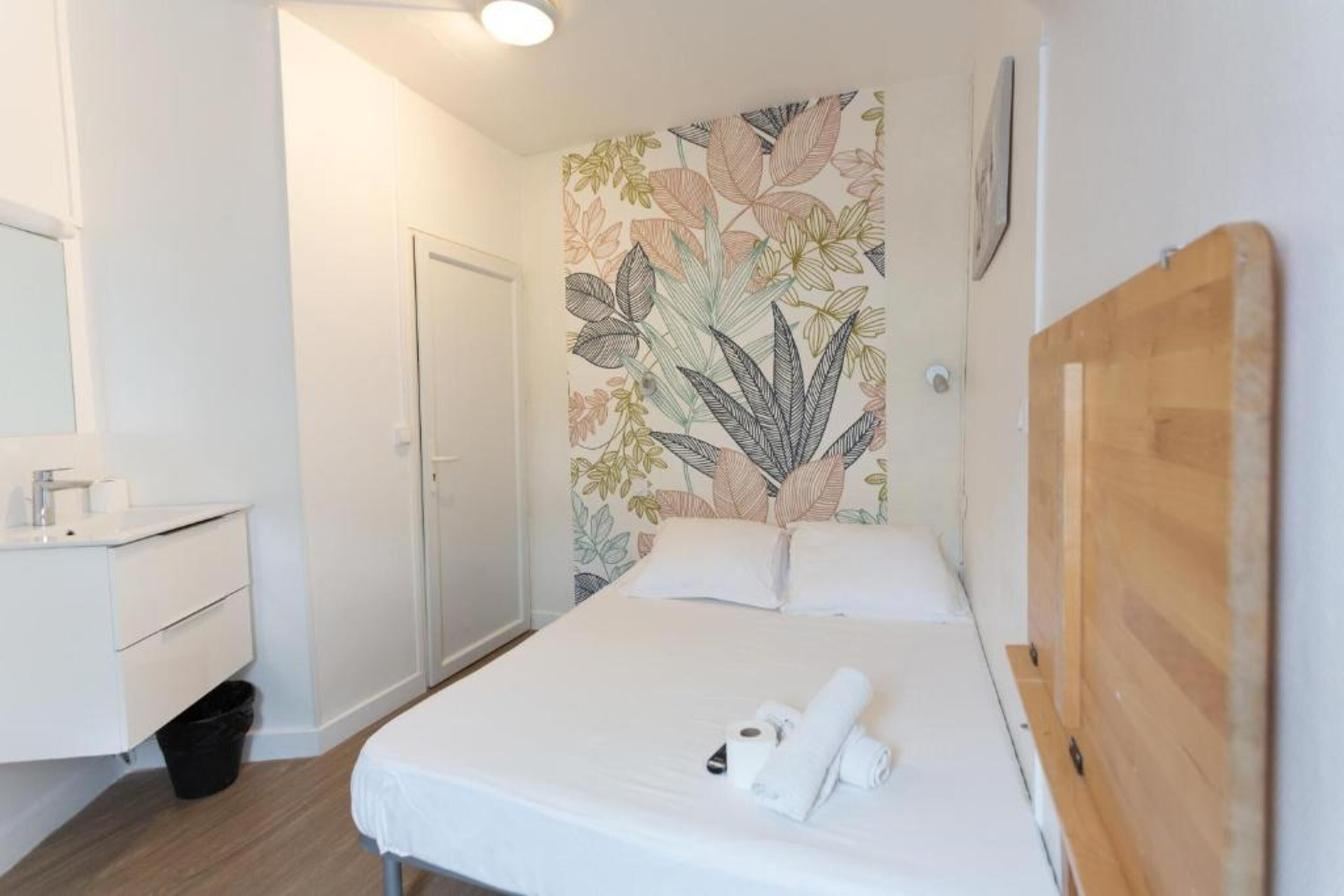 Et hotelværelse med en seng, et træhovedgærde, en hvid væg med et blomstermotiv, et hvidt natbord og et hvidt badeværelse med vask og spejl.