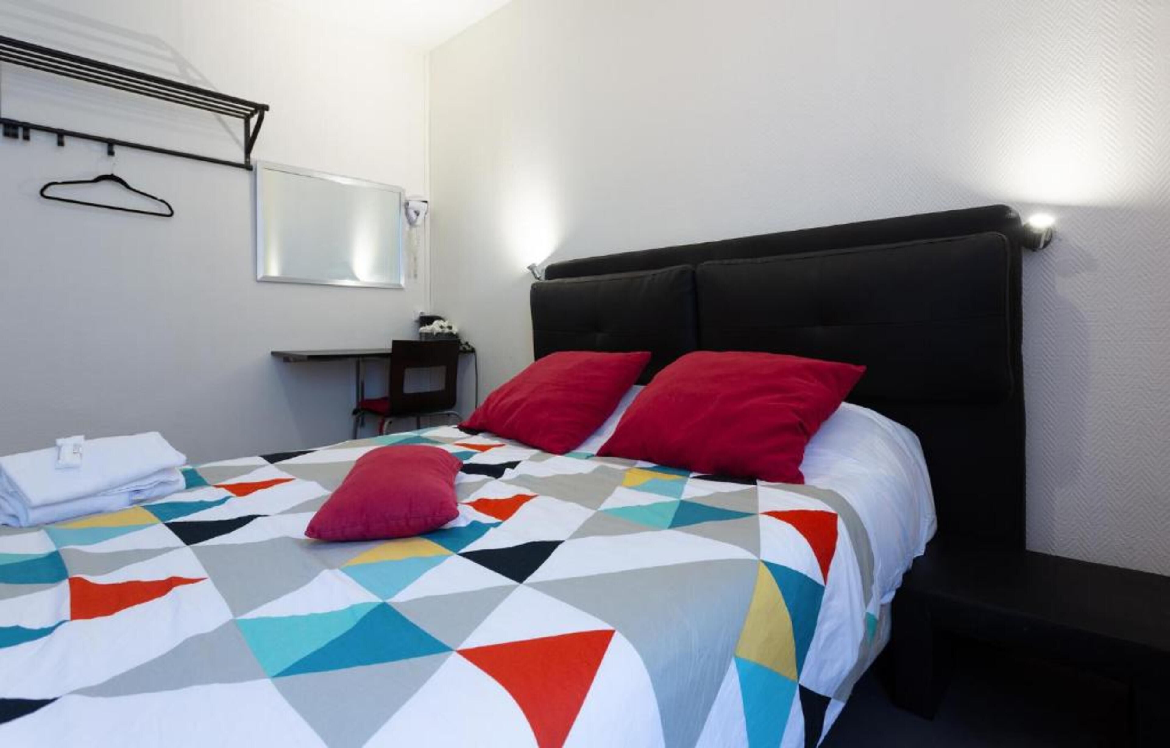A neatly made bed with a colorful geometric-patterned bedspread and two red pillows, a small bedside table with a lamp, and a wall-mounted hanger.