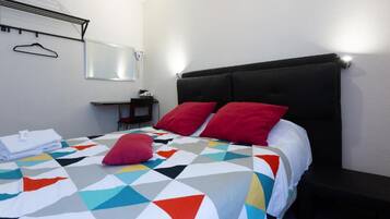 A neatly made bed with a colorful geometric-patterned bedspread and two red pillows, a small bedside table with a lamp, and a wall-mounted hanger.
