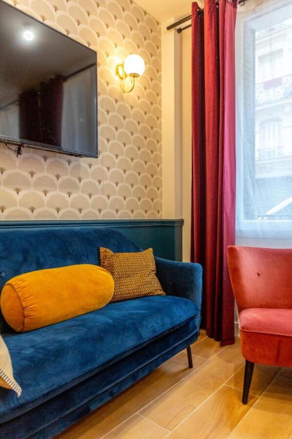 Una stanza con un divano blu, una poltrona rossa, una TV a schermo piatto montata a parete e una finestra con tende rosse.