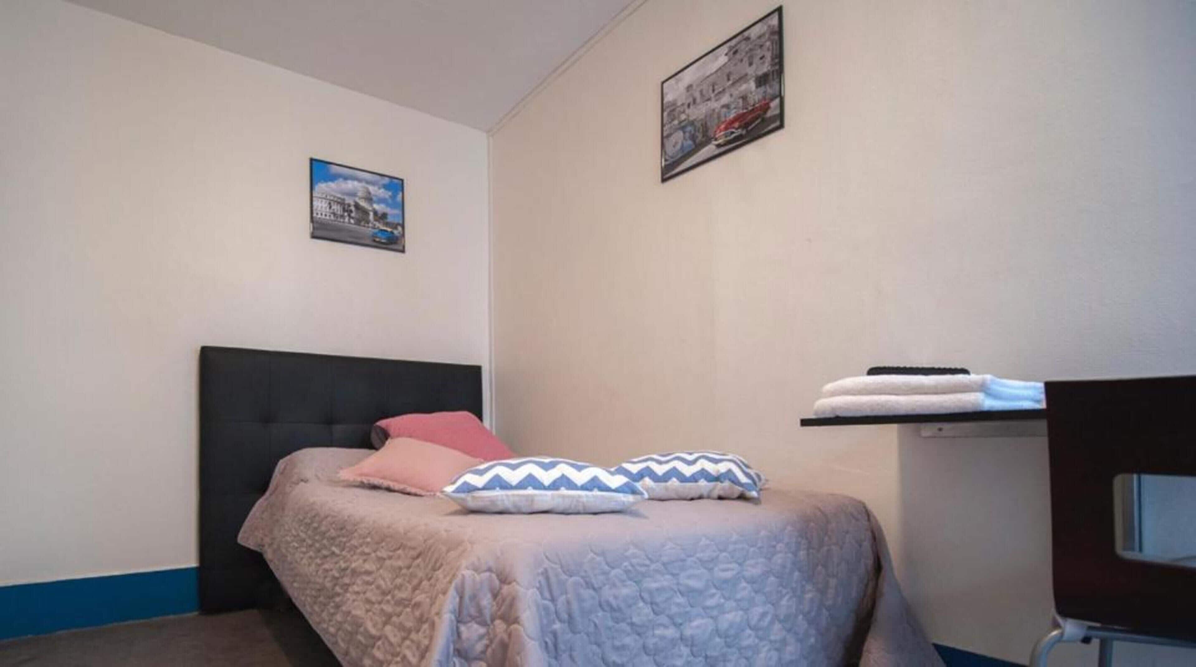 A bedroom with a bed, pillows, a headboard, a wall-mounted shelf with towels, and two framed pictures.