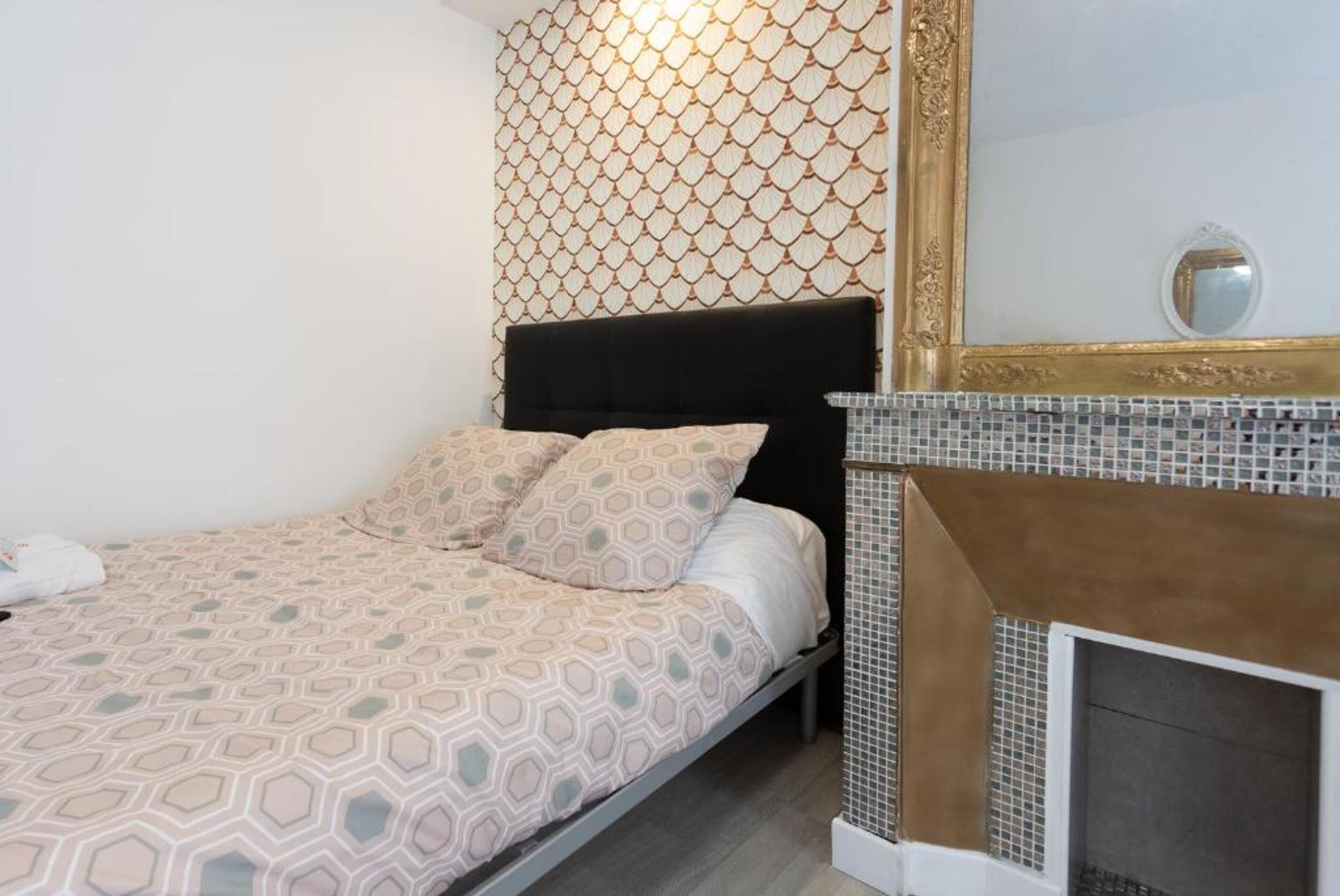 A bedroom with a patterned bedspread, a black headboard, a decorative mirror, and a tiled fireplace.