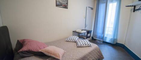 Un dormitorio con cama, almohadas, una silla, una mesita, televisor y ventana con cortinas.