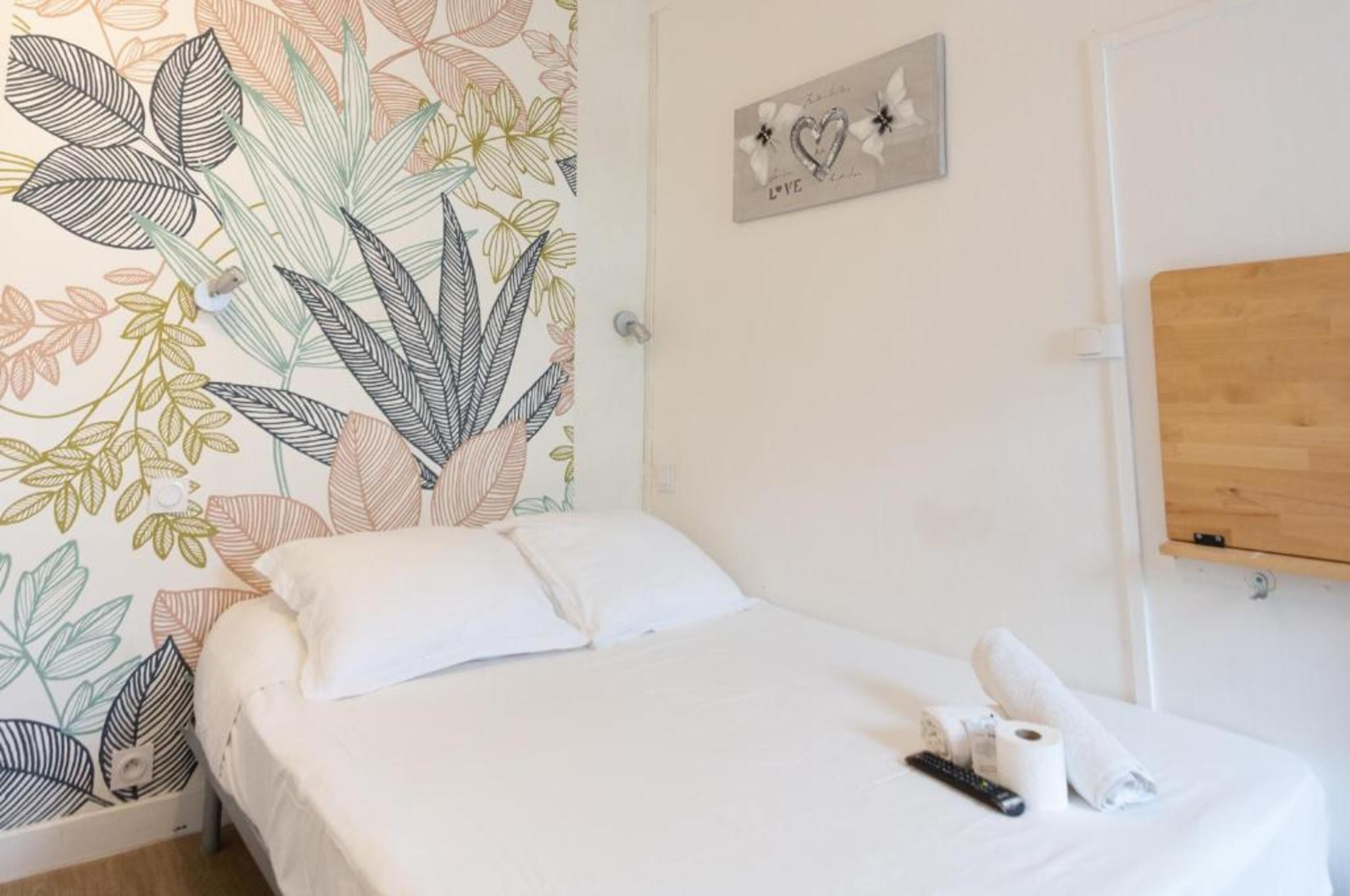 Habitació d'hotel amb un llit, una paret amb motius florals, una petita prestatgeria de fusta i un cartell amb cors i la paraula "LOVE".