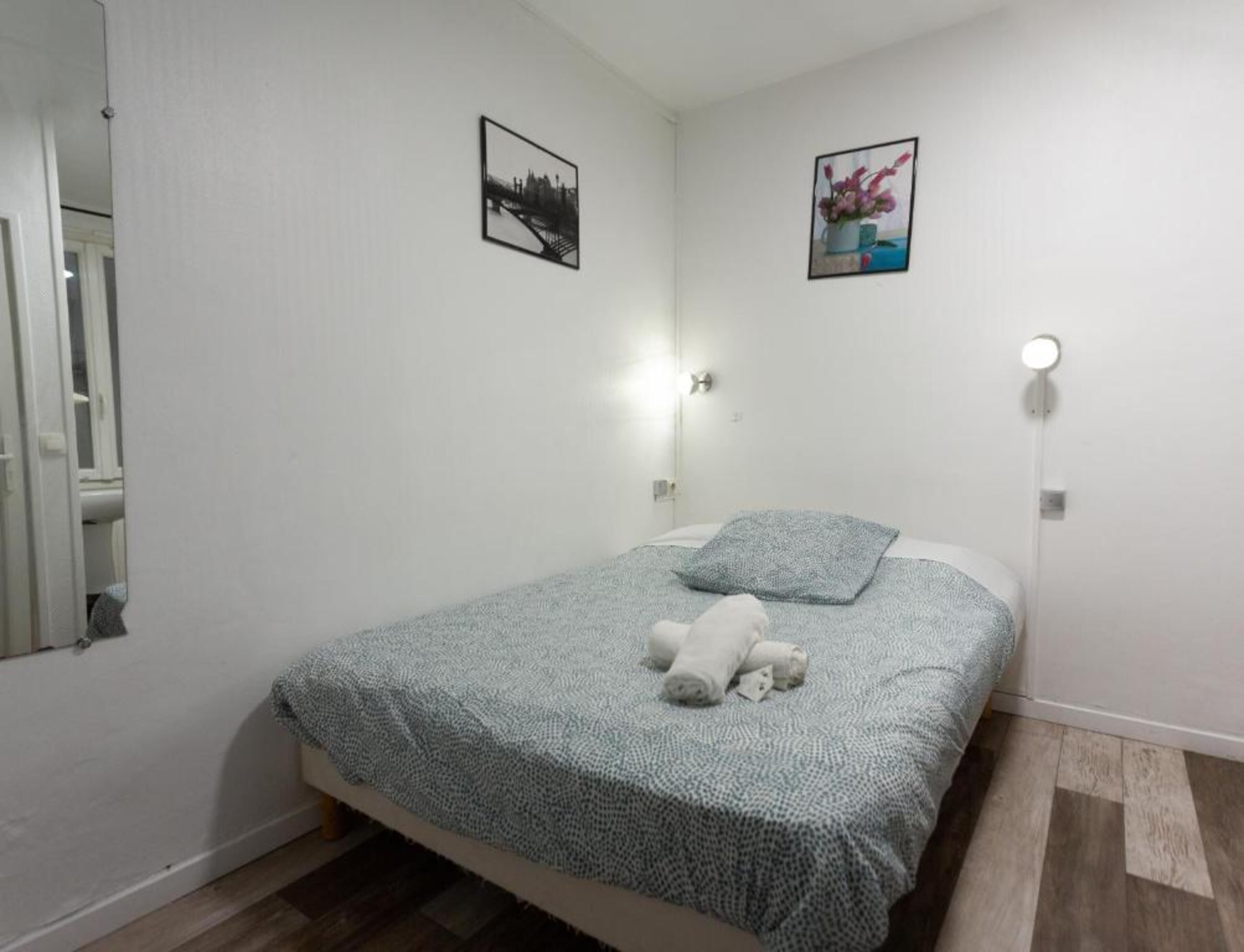 A single bed with a patterned bedspread, two wall-mounted light fixtures, and two framed pictures on the walls.