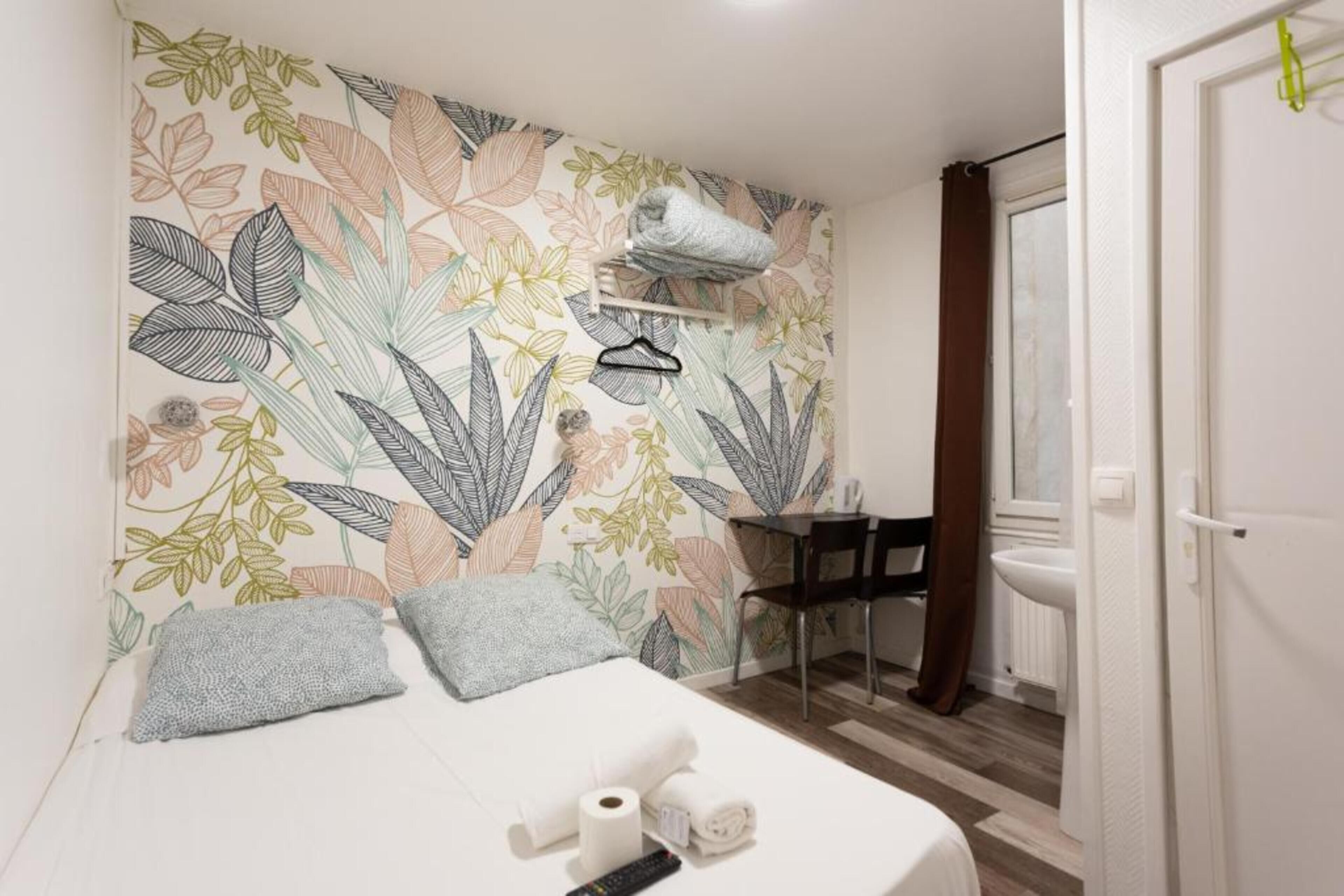 A hotel room with a bed, a wall with a floral pattern, a small table with a chair, and a bathroom visible through an open door.
