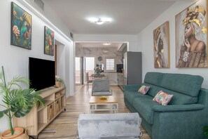 A modern living room with a flat-screen TV, a green sofa, and a dining area in the background.