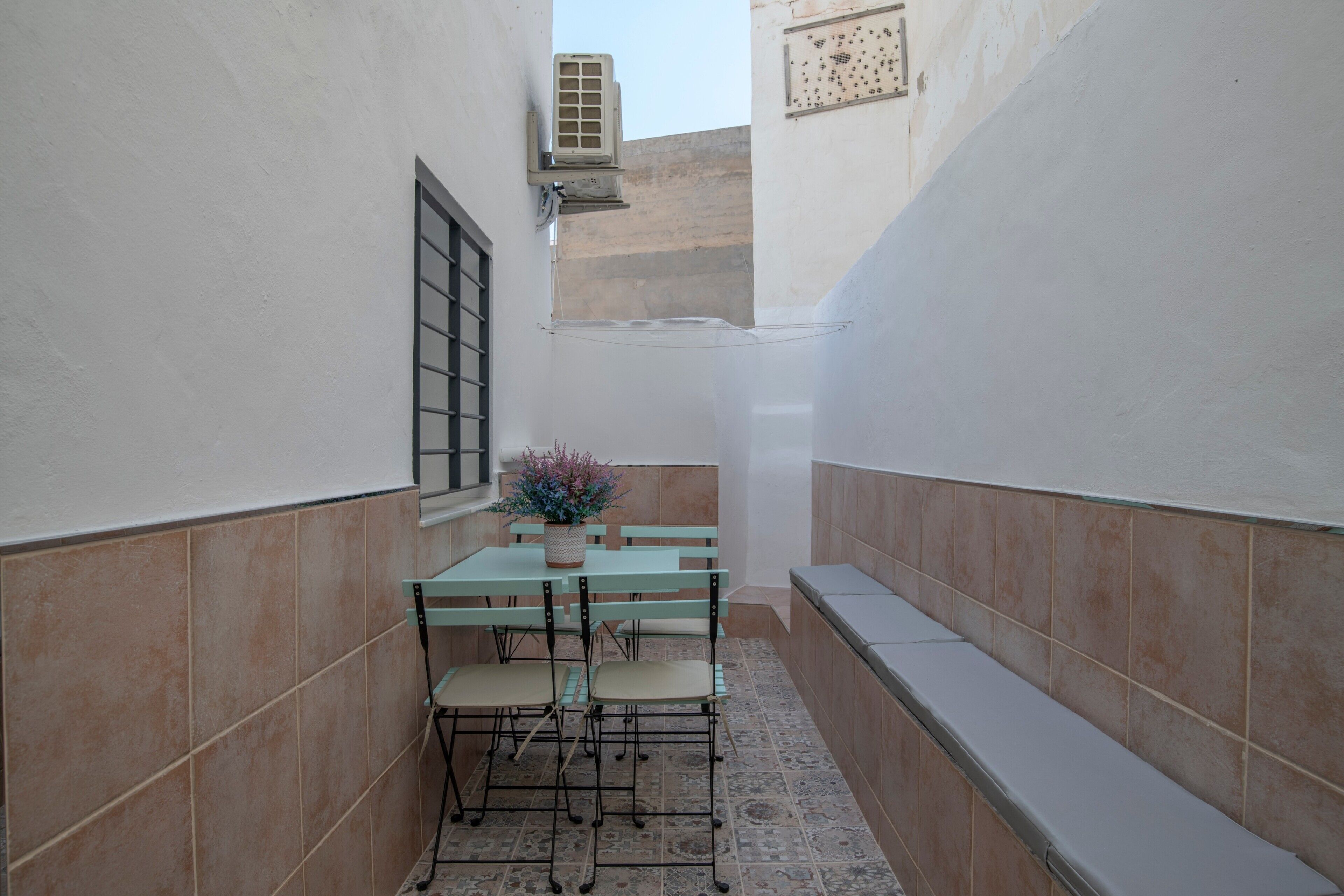 Un patio exterior estrecho con una mesa, sillas, una planta en maceta y una ventana con rejas.