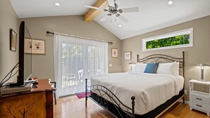 Un dormitorio con cama, escritorio, silla, ventilador de techo, ventana con vista a árboles y un cuadro en la pared.