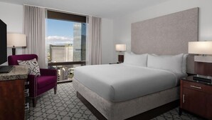 Ein Hotelzimmer mit einem großen Bett, einem Schreibtisch, einem Sessel und Blick auf eine Stadtkulisse durch ein großes Fenster.