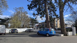 A parking area with several cars, including a blue hatchback, and a large tree in the foreground. - OYO Firs Lodge London Heathrow| Standard Double (Hounslow)