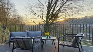 A balcony with a metal railing, two chairs, a small round table with a vase of flowers, and a view of a tree and distant landscape during sunset.