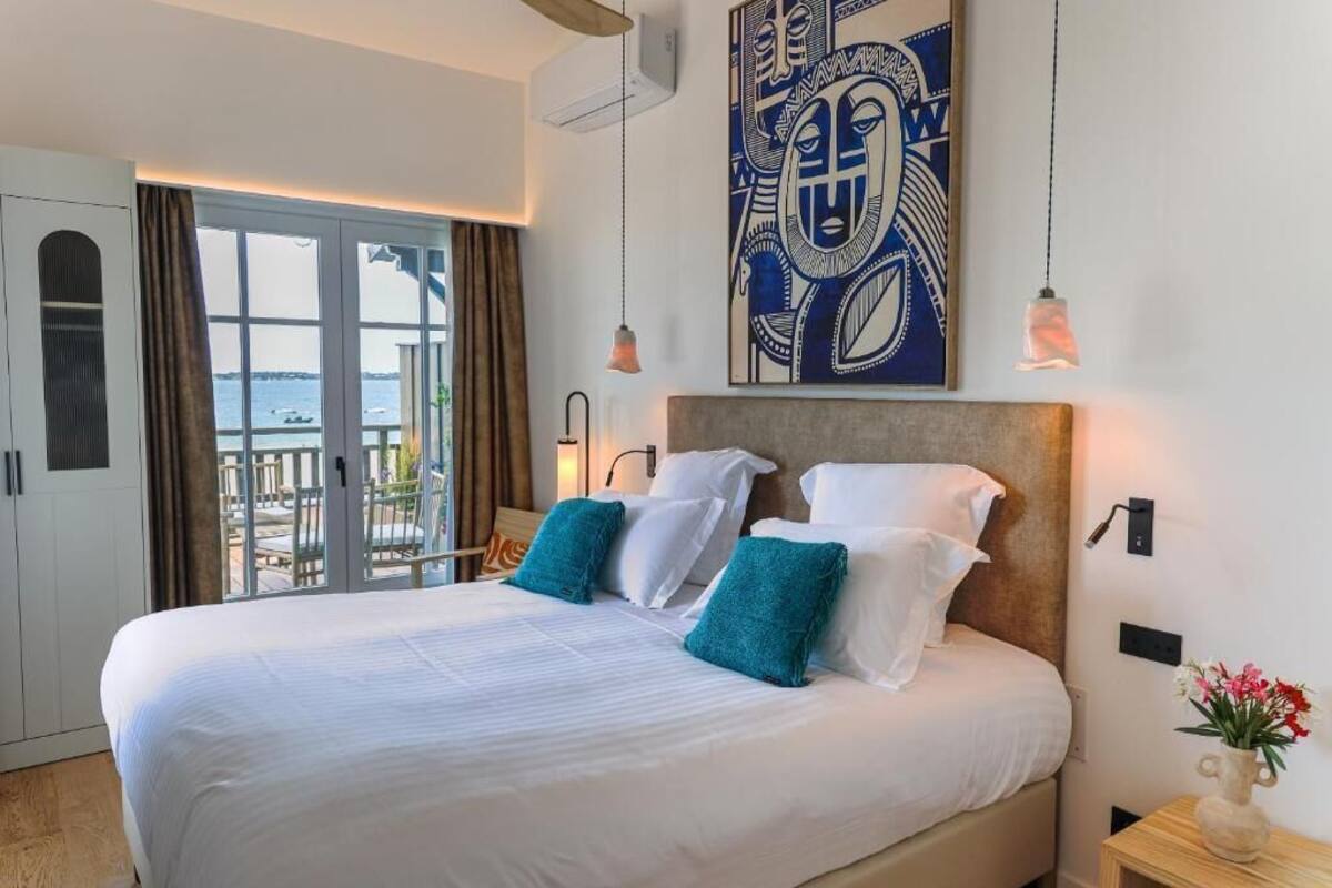 Habitación de hotel con una cama grande, vista al mar a través de una puerta corrediza, un cuadro abstracto moderno y un pequeño florero sobre la mesita de noche.
