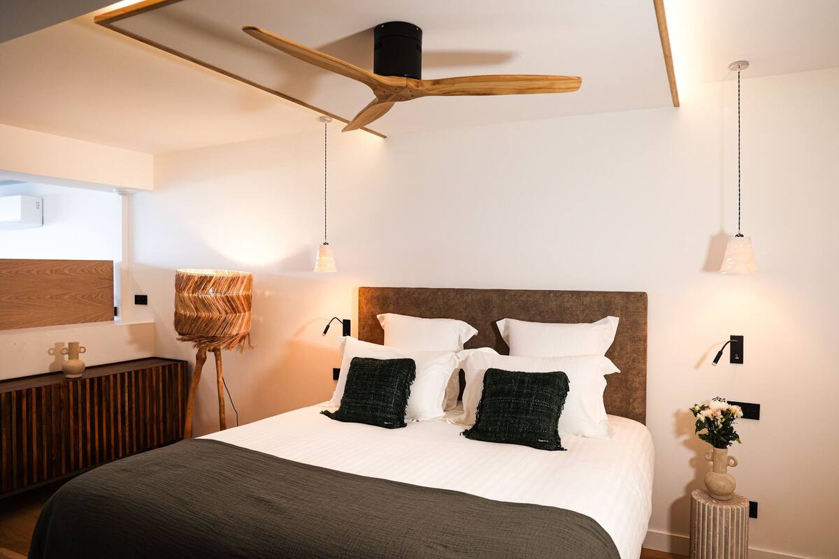 Una habitación de hotel moderna con una cama grande, un ventilador de techo de madera, una lámpara de noche y una pequeña mesa con un florero.