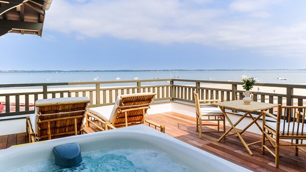 Une terrasse en bois avec un jacuzzi, des fauteuils en osier et une table agrémentée d’un vase de fleurs.